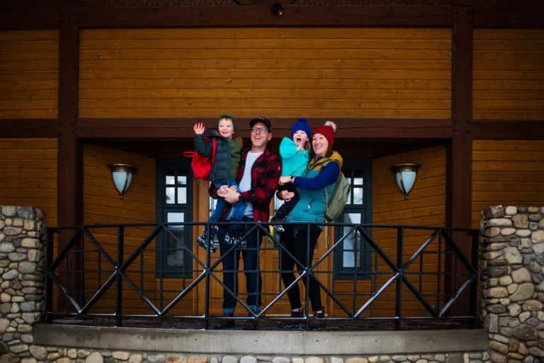 Smiling family on a wooden lodge balcony, dressed in winter gear with backpacks