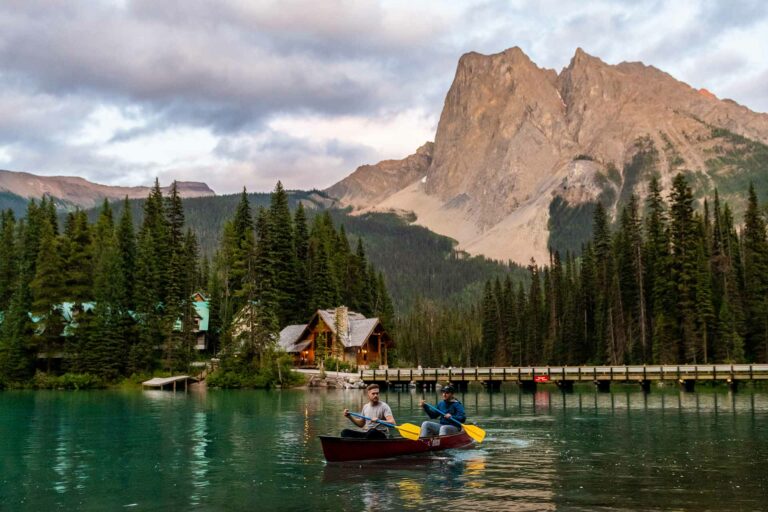 Two people canoeing on a tranquil lake, with a cozy lodge and mountain scenery in the background