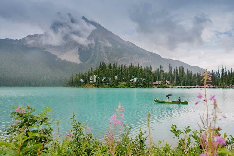 A couple paddles a canoe on a turquoise lake, with misty mountains and forested cabins in view