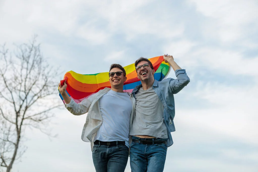 Celebrate Pride Month with CRMR - Sip with Pride, Give with Purpose! 1 Two smiling men in casual clothing hold a rainbow flag while walking outdoors