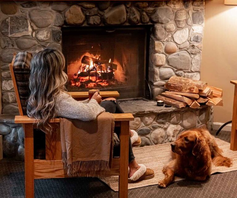 A woman relaxes by a stone fireplace with a golden retriever lying nearby.