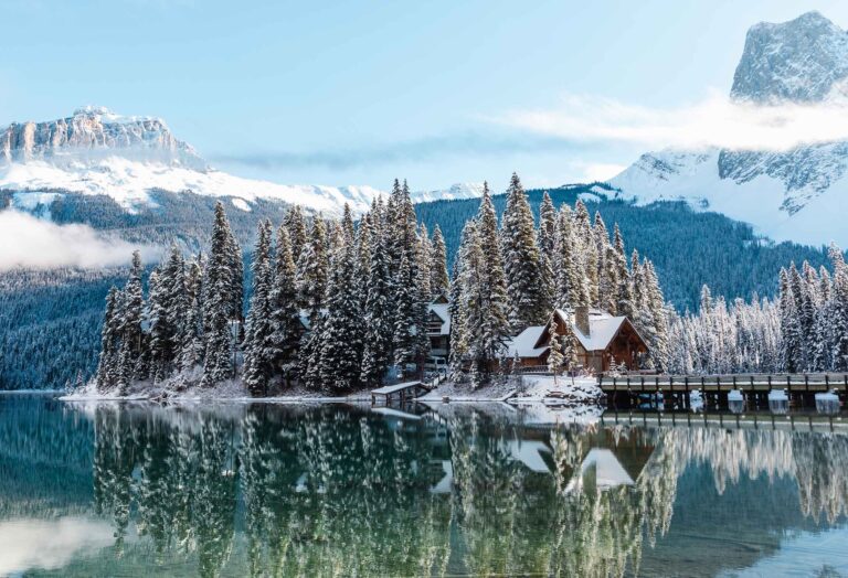 A snow-covered lodge sits by the tranquil Emerald Lake, surrounded by tall evergreen trees and mountains.
