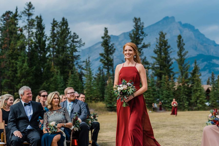 A bridesmaid in a flowing red dress walks down the aisle at an outdoor wedding with mountain views