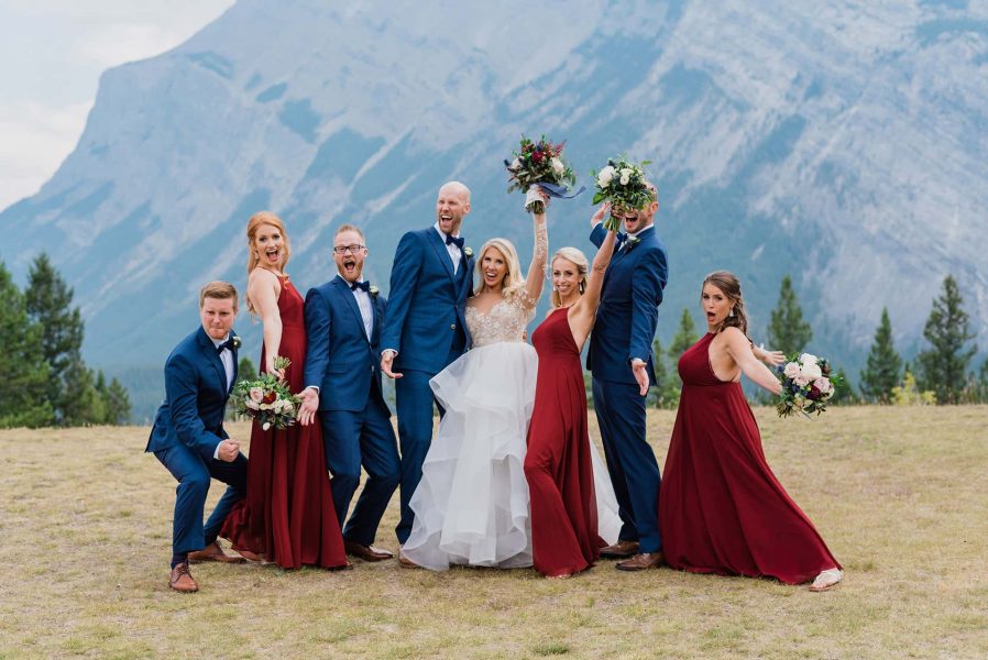 A joyful wedding party poses playfully outdoors with mountain scenery in the background