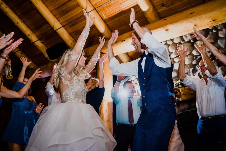 A bride and groom dance joyfully with guests at a rustic wedding reception in a log venue