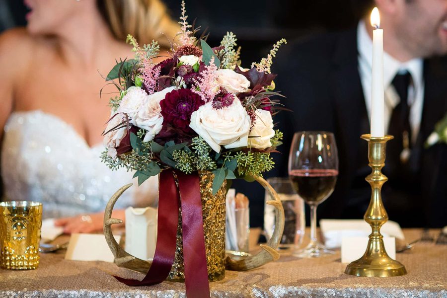 A floral centrepiece with white and burgundy roses in a gold vase with antlers, beside a candle